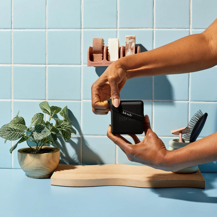 Hands fastening a black Kitsch travel case beneath shampoo and conditioner bars on a shower shelf.