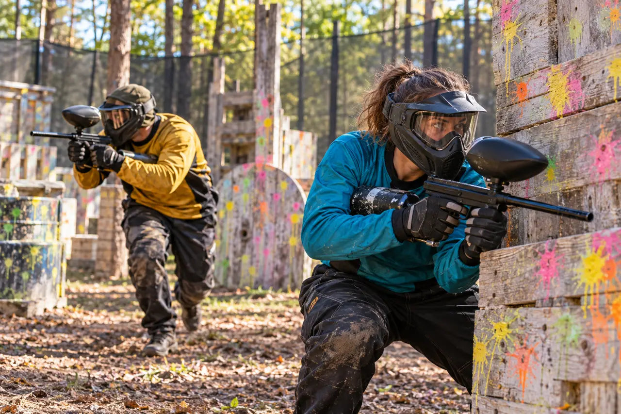 Two adults in full-face paintball masks and colorful jerseys playing behind wooden barriers on a designated field.
